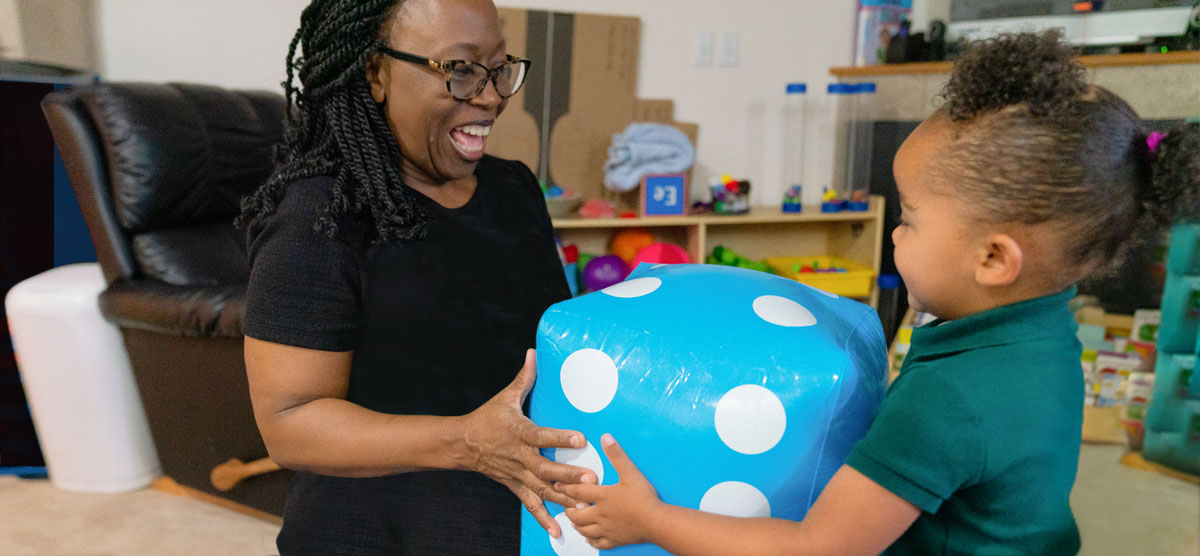 An adult and toddler are playing with large blow-up dice. The adult is laughing and smiling at the child,