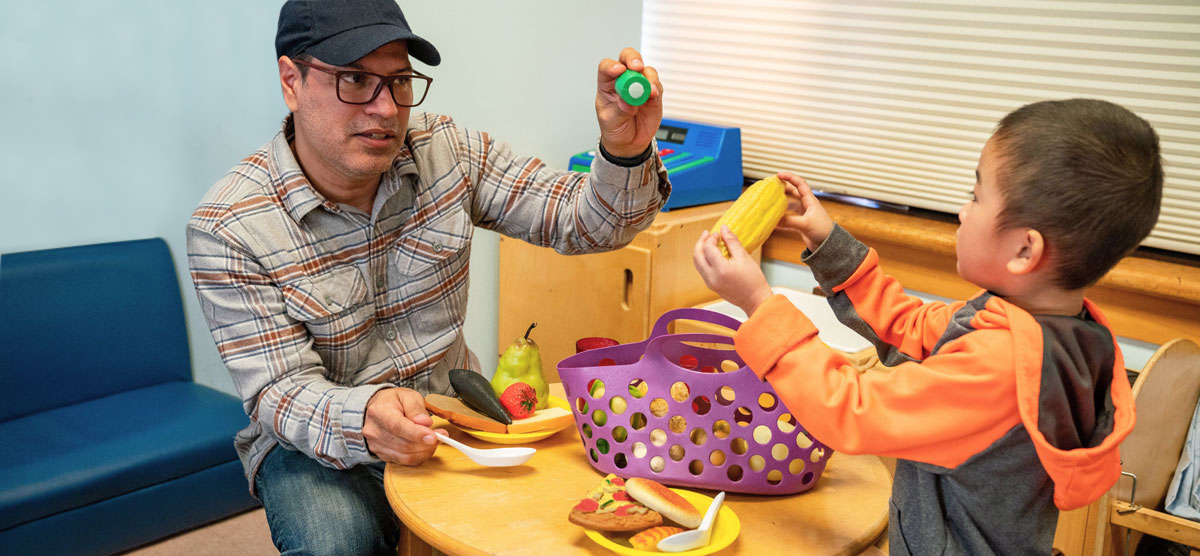 The educator and child are at the dramatic play area engaging with pretend food and dishes.