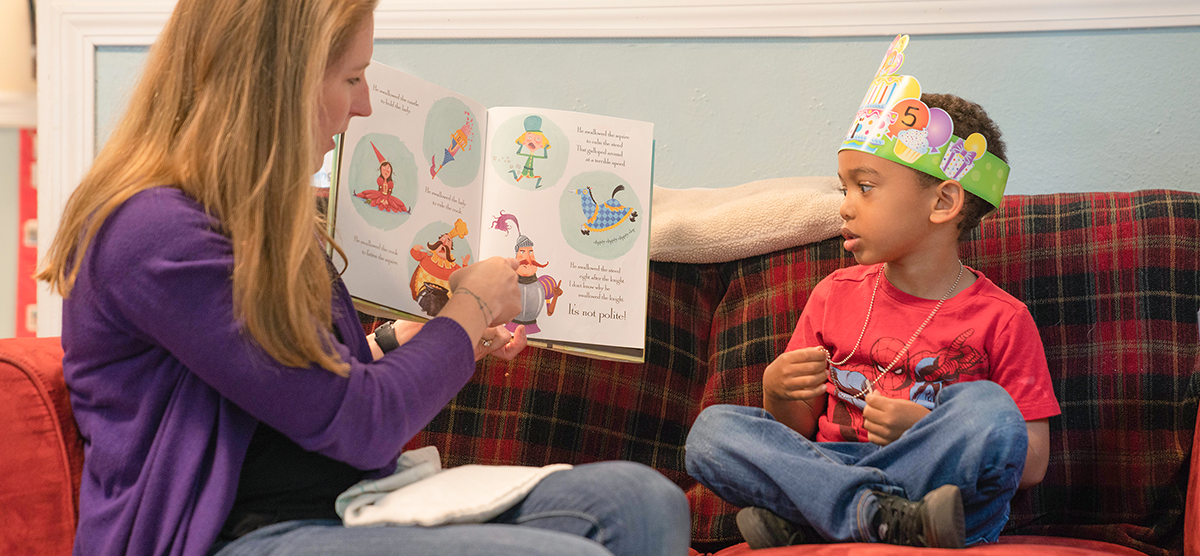 An educator is sitting on the couch with a child reading a book.