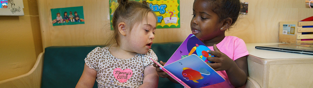 Two girls sit together on a couch reading board books together.