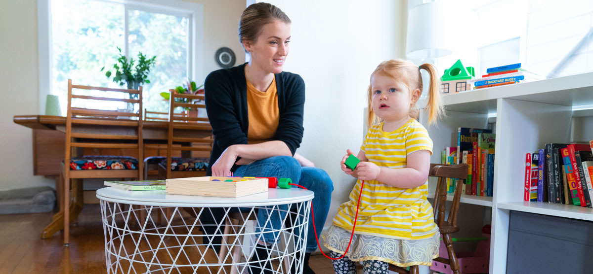 A child and her teacher sit at a child-sized table and put a series of blocks onto a string.