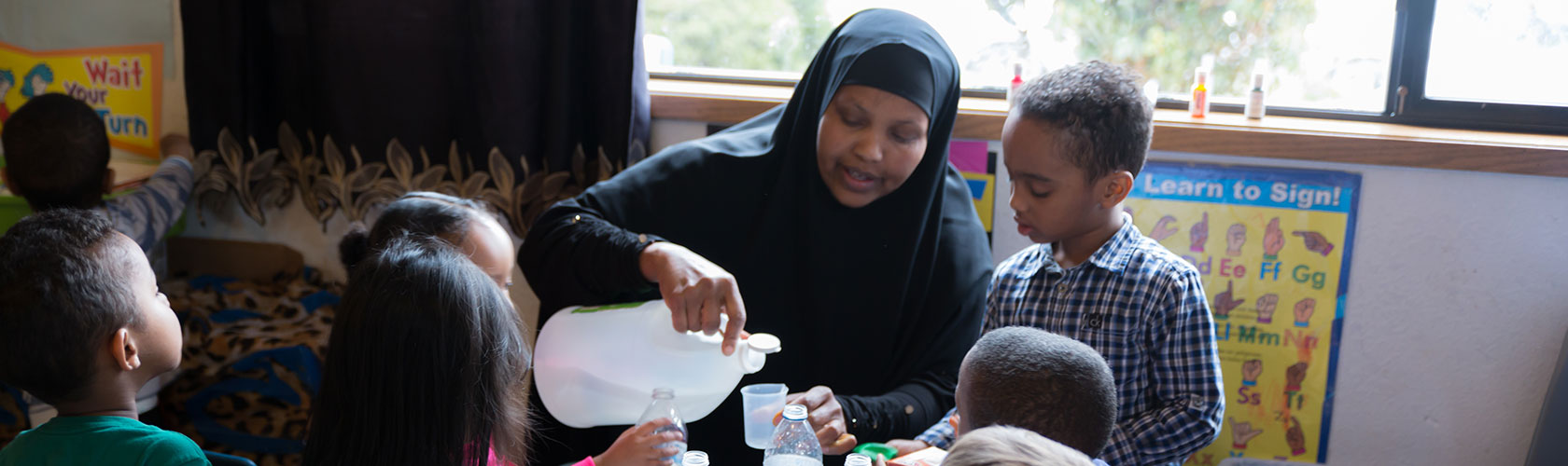 Caregiver pouring liquid into plastic cup while preschoolers surround her to watch.