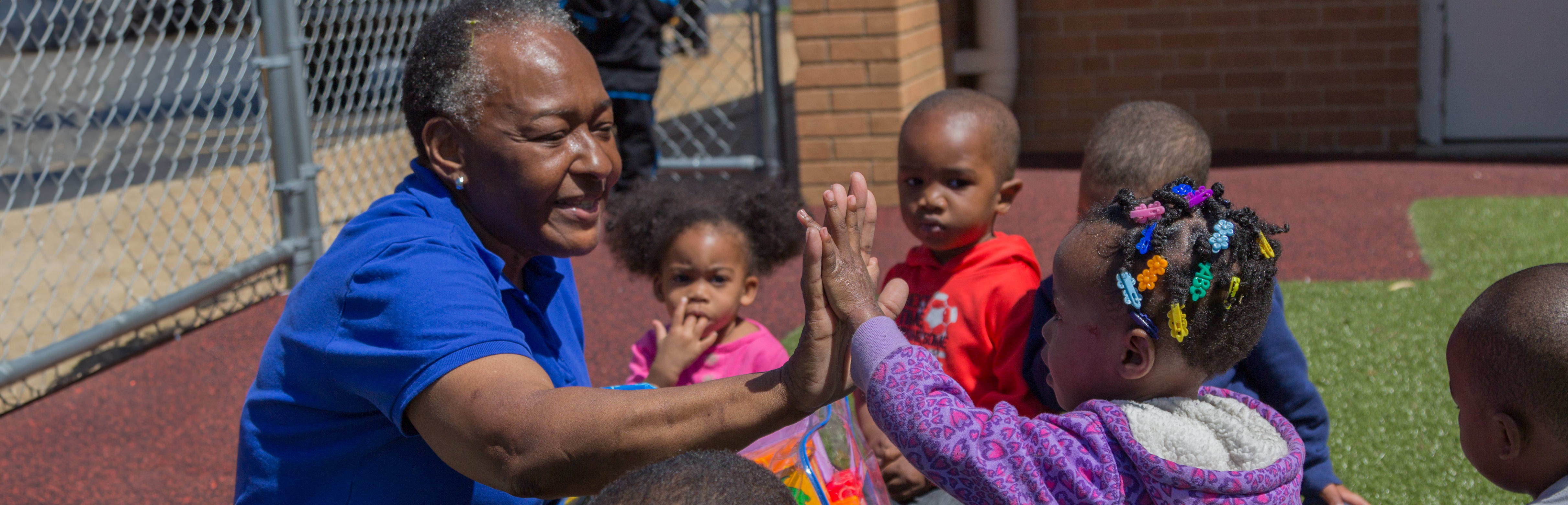 Caregiver high-fives a young child while other children gather around them outdoors.